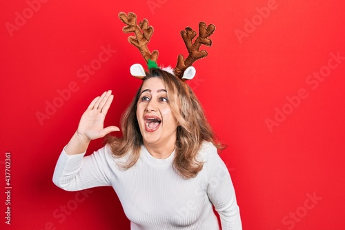 Middle age caucasian woman wearing cute christmas reindeer horns shouting and screaming loud to side with hand on mouth. communication concept.
