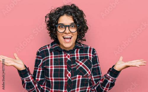 Young hispanic woman with curly hair wearing casual clothes and glasses celebrating victory with happy smile and winner expression with raised hands © Krakenimages.com