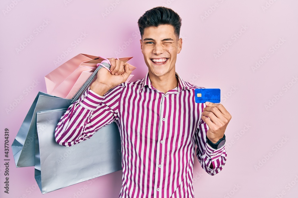 Young hispanic man holding shopping bags and credit card winking looking at the camera with sexy expression, cheerful and happy face. Young hispanic man holding shopping bags and credit card winking looking at the camera with sexy expression, cheerful and happy face.
