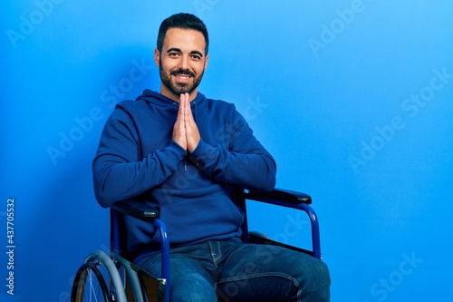 Handsome hispanic man with beard sitting on wheelchair praying with hands together asking for forgiveness smiling confident.