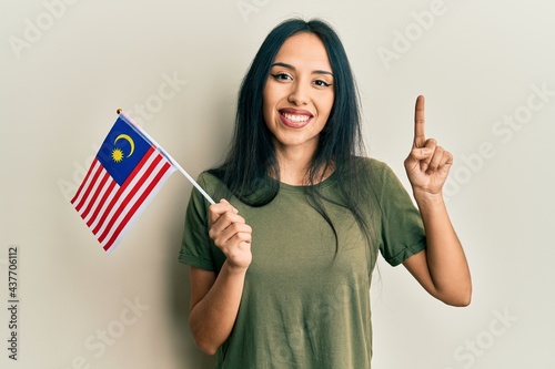 Young hispanic girl holding malaysia flag smiling with an idea or question pointing finger with happy face, number one