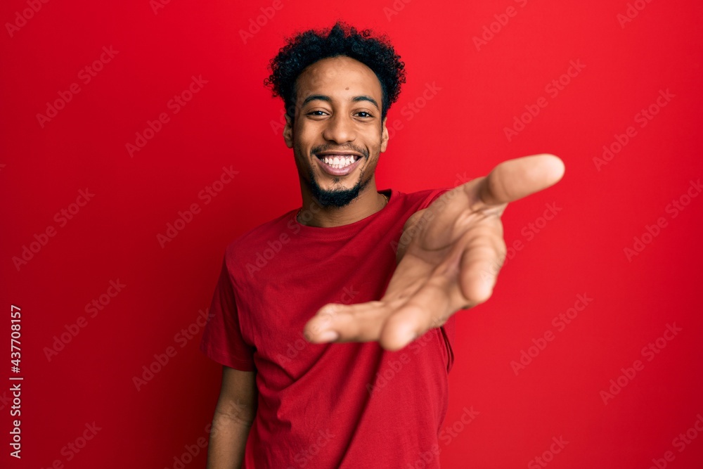 Young african american man with beard wearing casual red t shirt smiling friendly offering handshake as greeting and welcoming. successful business. Young african american man with beard wearing casual red t shirt smiling friendly offering handshake as greeting and welcoming. successful business.