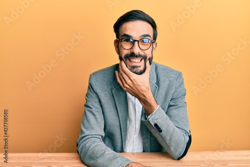 Young hispanic man working at the office looking confident at the camera smiling with crossed arms and hand raised on chin. thinking positive.