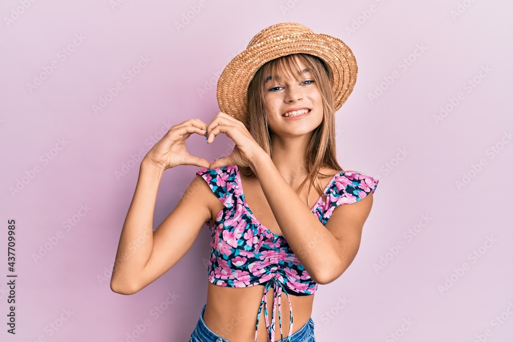 Teenager caucasian girl wearing summer hat smiling in love doing heart symbol shape with hands. romantic concept.