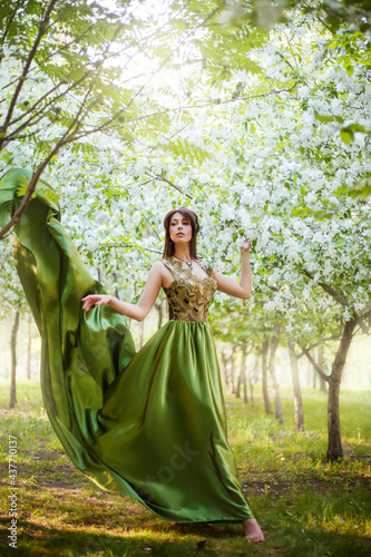 A beautiful young woman in a green dress with a large train among the blooming apple trees.