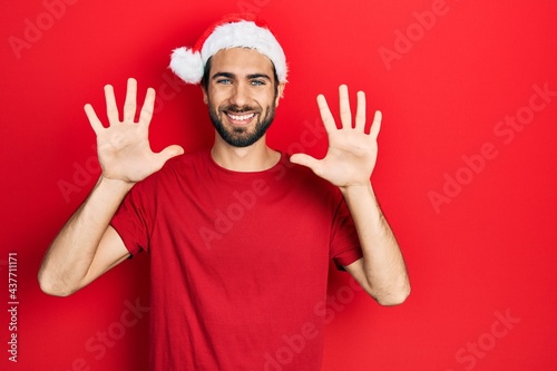 Young hispanic man wearing christmas hat showing and pointing up with fingers number ten while smiling confident and happy.