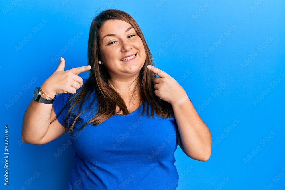 Beautiful brunette plus size woman wearing casual blue t shirt smiling cheerful showing and pointing with fingers teeth and mouth. dental health concept. Beautiful brunette plus size woman wearing casual blue t shirt smiling cheerful showing and pointing with fingers teeth and mouth. dental health concept.