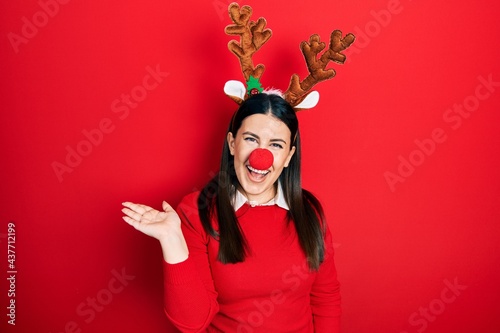 Young hispanic woman wearing deer christmas hat and red nose smiling cheerful presenting and pointing with palm of hand looking at the camera.