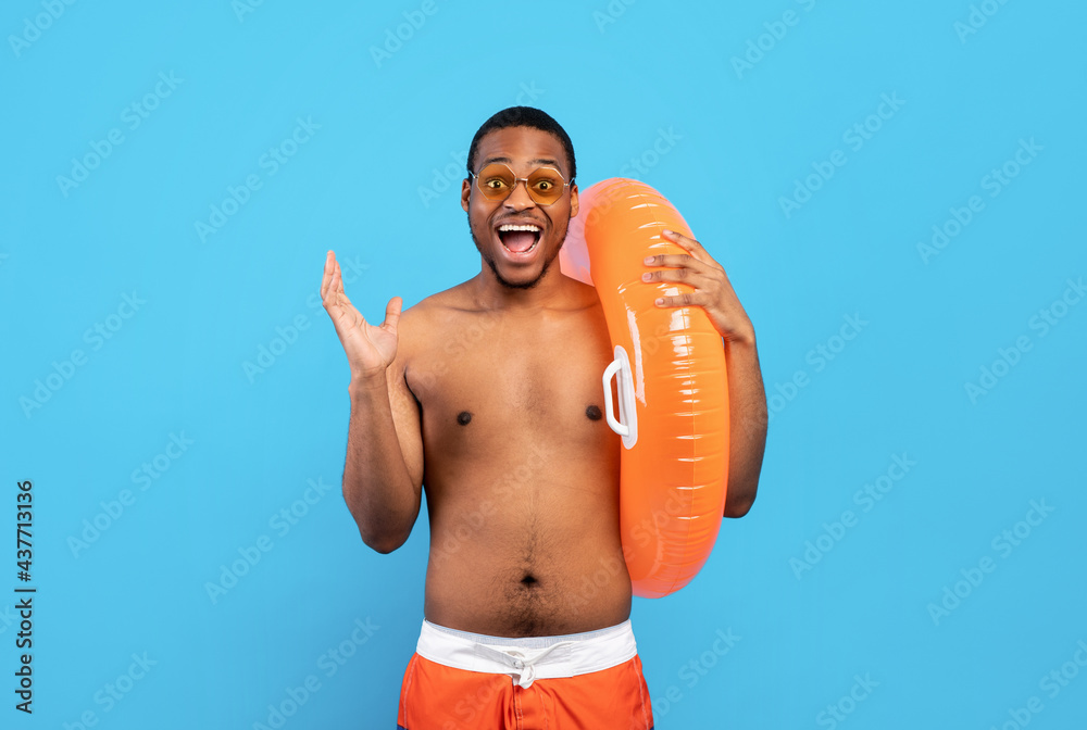 Emotional black guy in swimwear holding inflatable ring, shouting WOW, having great summer vacation, blue background