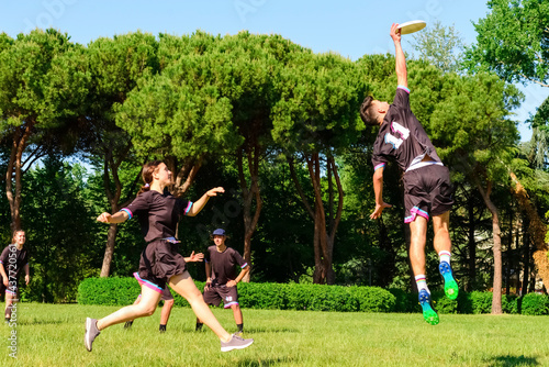 Group of young teenagers people in team wear playing a frisbee game in park oudoors. jumping man catch a frisbee to a teammate in an ultimate frisbee match. milennials friends outside in a garden photo