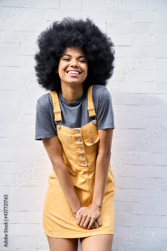 Portrait of joyful stylish black African American teenage hipster girl wearing trendy yellow sundress with afro hair standing against white bricks wall background smiling looking at camera.