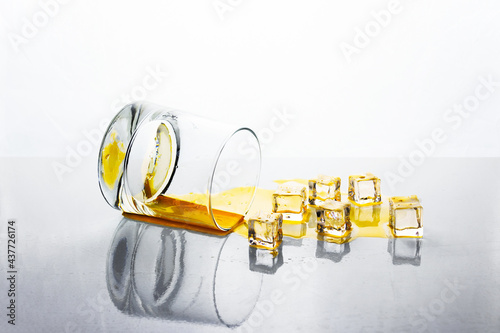 Knocked over glass of golden alcohol with ice cubes on white reflective surface, whiskey or cognac. photo