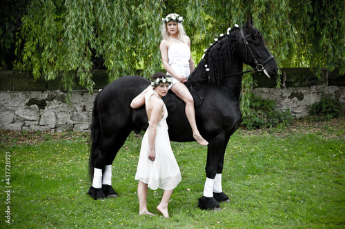 Fotografie Two young woman in white dresses with a black shire horse