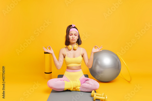 Beautiful relaxed Asian woman in sportswear practices yoga which brings energy sits in lotus position being in harmony with body surrounded by sport equipment isolated over yellow background
