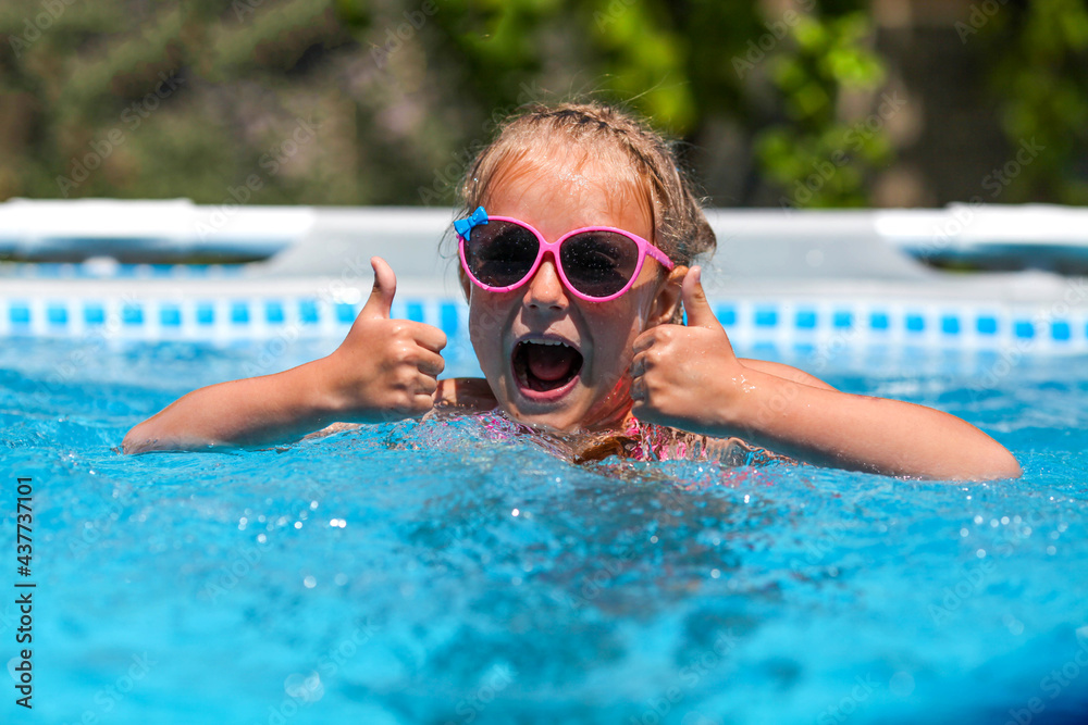 Cheerful little girl smiling in sunglasses in the pool on a sunny day. The child showing thumbs up like gesture looking camera . Summer vacation