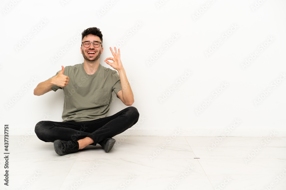 Young caucasian man sitting on the floor isolated on white background showing ok sign and thumb up gesture Young caucasian man sitting on the floor isolated on white background showing ok sign and thumb up gesture