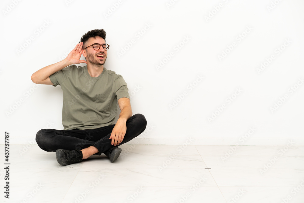 Young caucasian man sitting on the floor isolated on white background listening to something by putting hand on the ear Young caucasian man sitting on the floor isolated on white background listening to something by putting hand on the ear