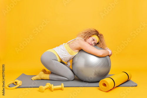 Fitness and aerobics concept. Tired curly haired female gymnast leans at fitball dressed in activewear uses dumbbells for exercising poses on fitness mat isolated over yellow studio background. photo