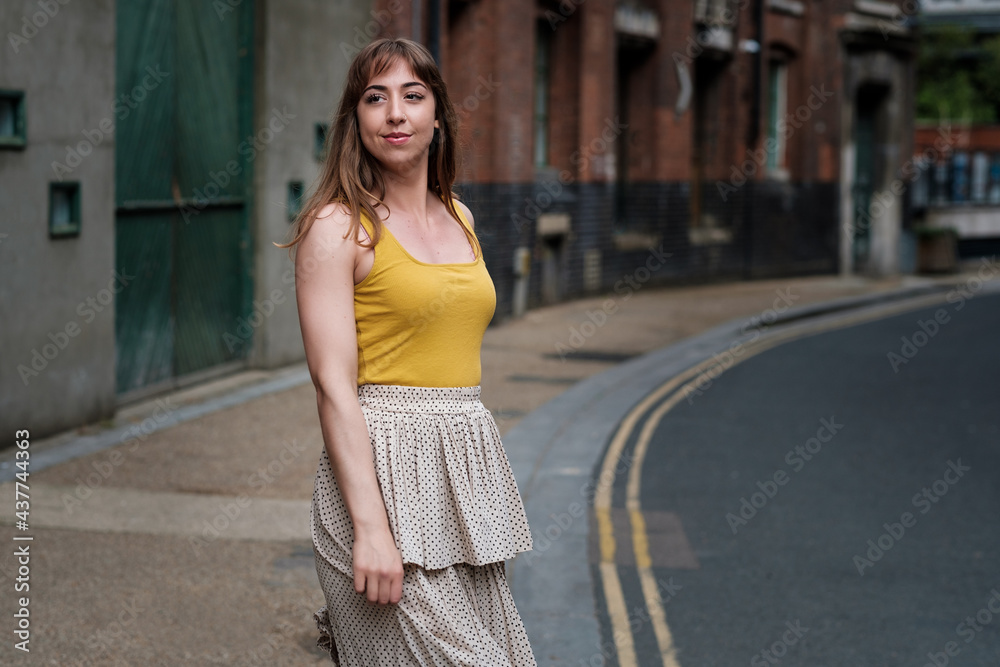 Young woman in a yellow t-shirt and white skirt posing in a narrow street.