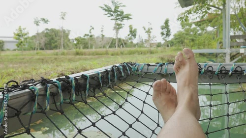 4K video of man's bare feet laying down on crandall net bed above water in farm field of the rural area in asian country (Thailand) shows relaxing, enjoyment and happiness for vacation in nature. photo