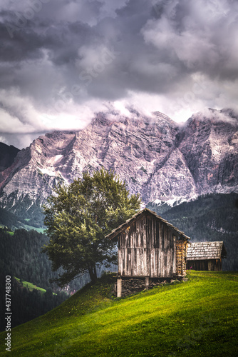 Wooden cottages near a tree on a grassy hill with mountains in the background and dark cloudy sky in Dolomites, Italy