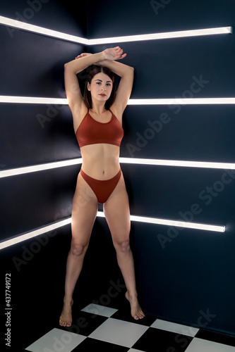 Portrait of beautiful caucasian brunette woman in maroon coloured swimsuit lingerie standing against dark wall with fluorescent lamps. Selective focus. Beauty theme.