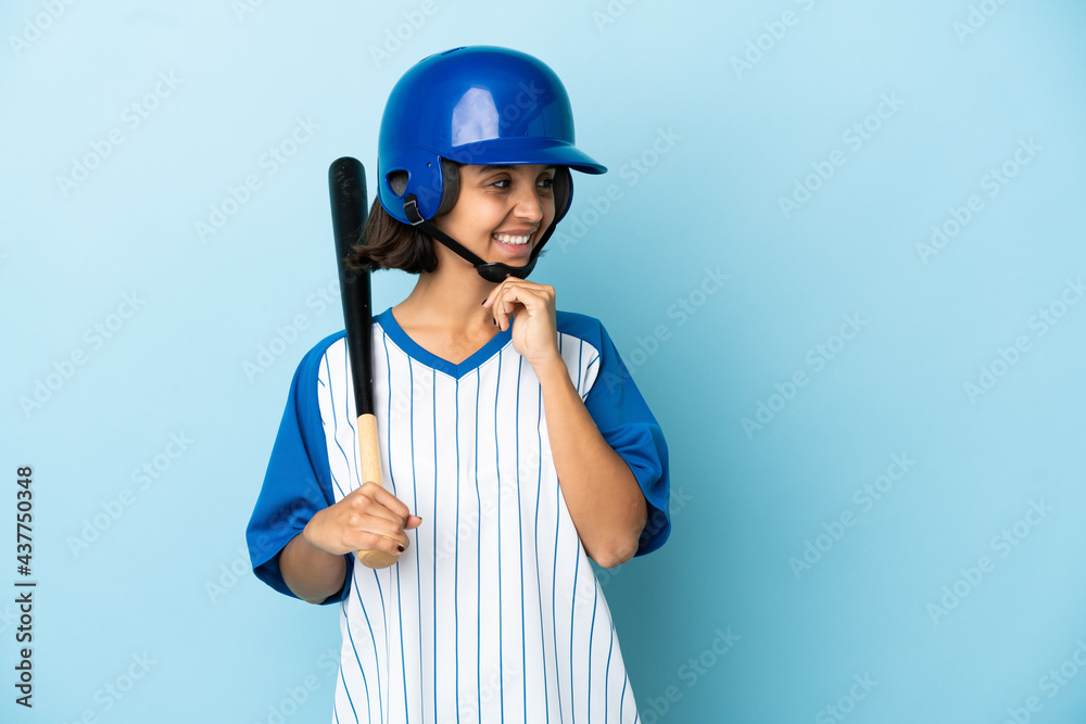 Baseball mixed race player woman with helmet and bat isolated on blue background looking to the side and smiling