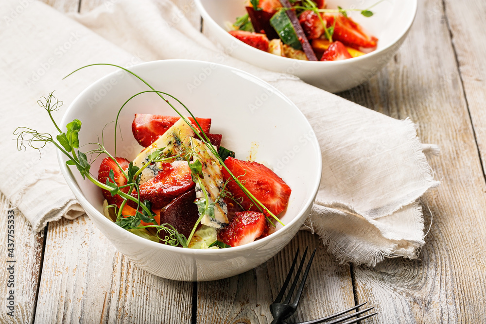Close-up delicious summer salad. Salad with fresh strawberries, baked beets, yellow tomatoes, dor blue cheese and herbs in a white bowl on a light rustic background. Summerfood. Close-up delicious summer salad. Salad with fresh strawberries, baked beets, yellow tomatoes, dor blue cheese and herbs in a white bowl on a light rustic background. Summerfood.