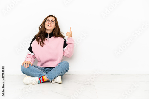 Young caucasian woman sitting on the floor isolated on white background pointing up and surprised © luismolinero