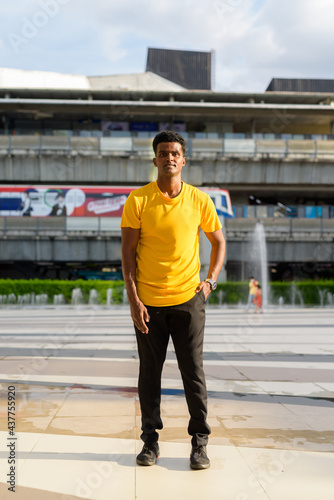 Full length portrait of handsome black African man wearing yellow t-shirt outdoors in city during summer