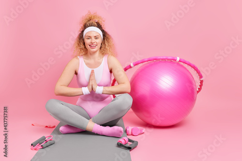 Photo of relaxed woman with curly hair keeps palms pressed together legs crossed practices yoga has fitness training surrounded by sport equipment does sport exercises at home. Active lifestyle