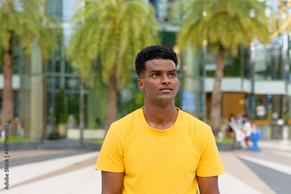 Portrait of handsome black African man wearing yellow t-shirt outdoors in city during summer while thinking