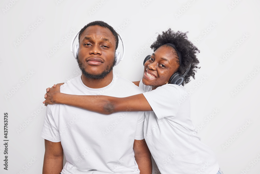 Cheerful dark skinned woman embraces her best male friend wear casual white t shirts wireless headphones have friendly relationships enjoy spending time together. Afro American couple indoor Cheerful dark skinned woman embraces her best male friend wear casual white t shirts wireless headphones have friendly relationships enjoy spending time together. Afro American couple indoor