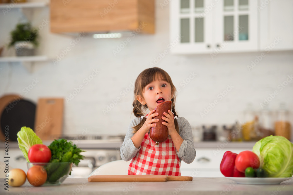 Cute little girl with pigtails in a checkered culinary apron eating sausage in the kitchen at the table