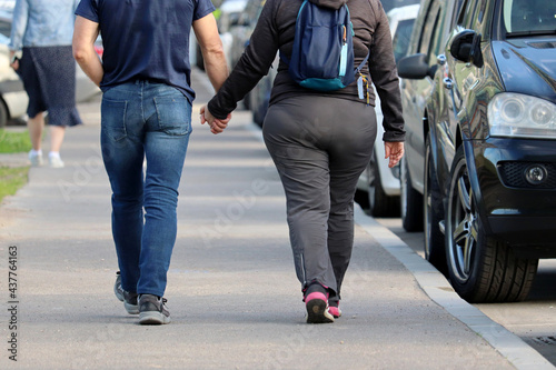 Fat woman in sportswear and slim man in jeans walking along a city street with parked cars. Couple in love holding hands, summer fashion