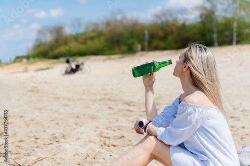 Attractive blonde woman is holding a bottle of cold beer in summer on beach near the lake.