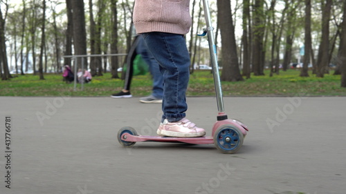 A girl in the park rides a three-wheeled scooter on the asphalt