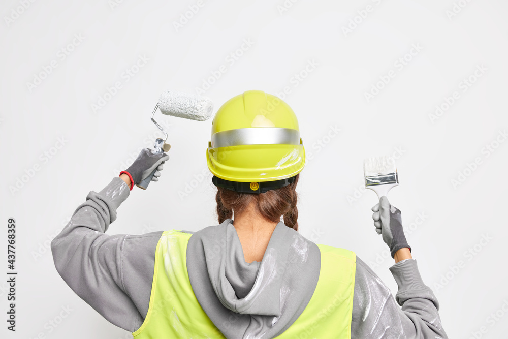 Horizontal shot of experienced female builder poses back to camera paints wall with painting roller and brush wears helmet and uniform busy doing apartment decoration. Skillful woman painter Horizontal shot of experienced female builder poses back to camera paints wall with painting roller and brush wears helmet and uniform busy doing apartment decoration. Skillful woman painter