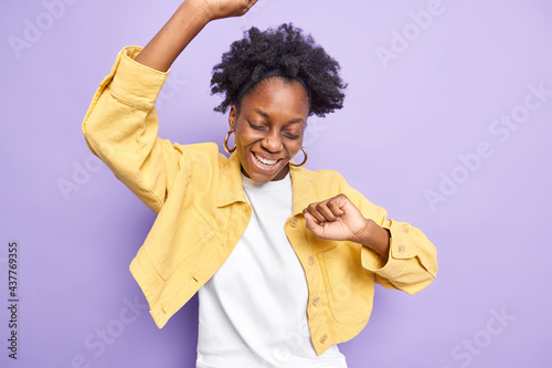 Upbeat dark skinned curly haired woman cheers and keeps arms raised up feels like lcky winner celebrates victory makes triumph dance smiles upbeat moves against purple background dressed casually