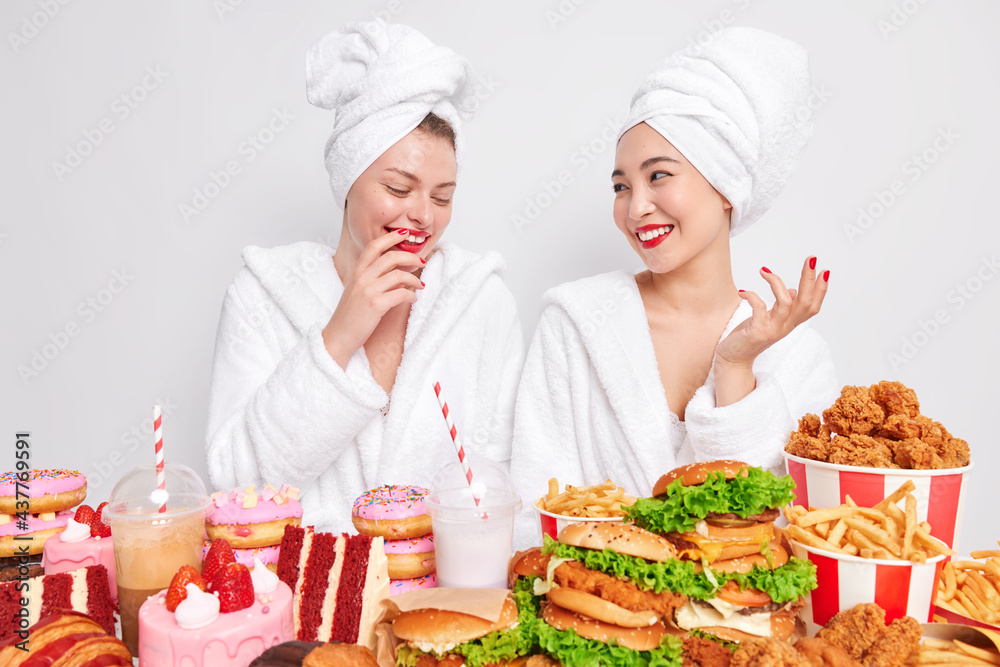 Two happy mixed race women have fun after taking shower stand next to each other wears comfortable bathrobe pose near table full of fast food eats cheat meal do not care about proper nutrition. Two happy mixed race women have fun after taking shower stand next to each other wears comfortable bathrobe pose near table full of fast food eats cheat meal do not care about proper nutrition.