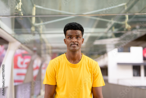 Portrait of handsome black African man wearing yellow t-shirt outdoors in city during summer while looking at camera at footbridge