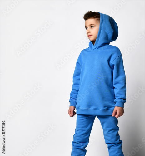 stylish boy in a blue sports sweatshirt with a hood and pants is standing with a hood over his head, pointing somewhere to the side. Street style accessories trends. look away