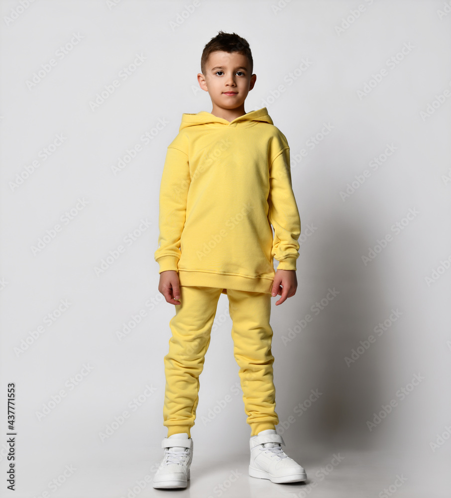 Stylish brunet boy child in a sports sweatshirt with a hood and pants stands with a hood over his head, hands in his pockets. The fashionable student is ready for school. Stylish brunet boy child in a sports sweatshirt with a hood and pants stands with a hood over his head, hands in his pockets. The fashionable student is ready for school.
