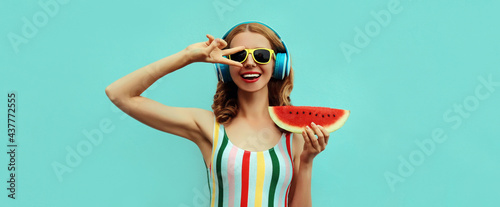 Summer portrait of cheerful happy smiling young woman in headphones listening to music with juicy slice of watermelon on a blue background