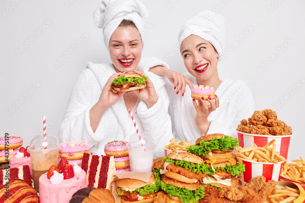 Happy women enjoy cheat meal eats tasty burgers cakes and doughnuts being addicted to fast food have unhealthy nutrition stand near table full of tasty appetizing snacks harmful for your health