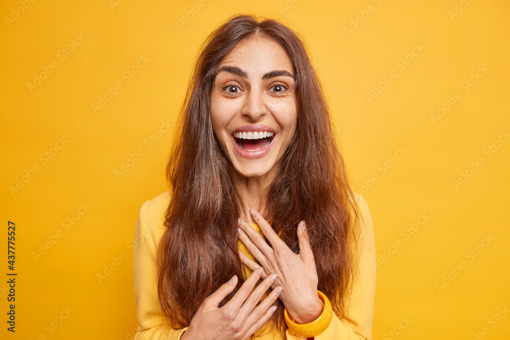 Excited happy European woman with natural long hair smiles broadly hears excellent news smiles broadly shows white teeth feels very glad poses against vivid yellow background. Human emotions Excited happy European woman with natural long hair smiles broadly hears excellent news smiles broadly shows white teeth feels very glad poses against vivid yellow background. Human emotions