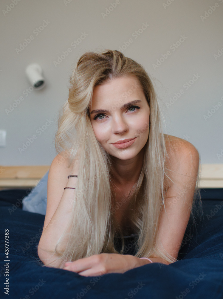Wonderful day happy smiling blonde young blue-eyed woman having fun on the sofa staring into camera holding her head dressed blue denim jeans grey bra Wonderful day happy smiling blonde young blue-eyed woman having fun on the sofa staring into camera holding her head dressed blue denim jeans grey bra