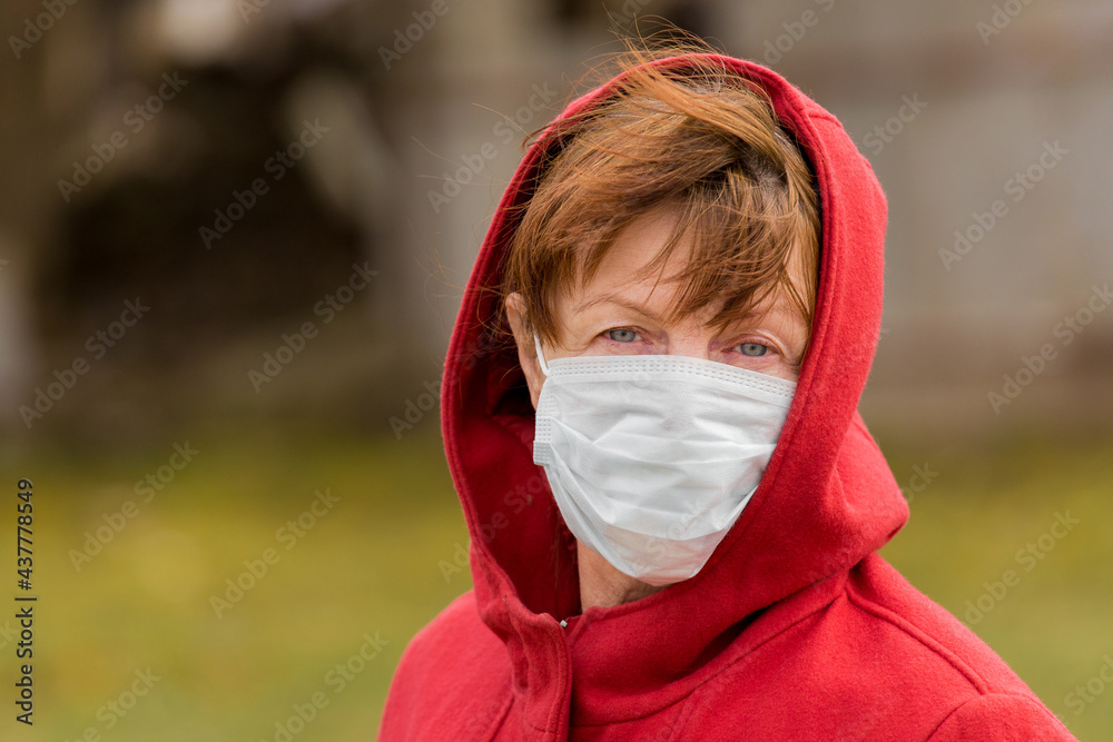 An elderly woman with brown hair and blue eyes in a red coat and hood in a protective safe medical mask, close-up portrait An elderly woman with brown hair and blue eyes in a red coat and hood in a protective safe medical mask, close-up portrait