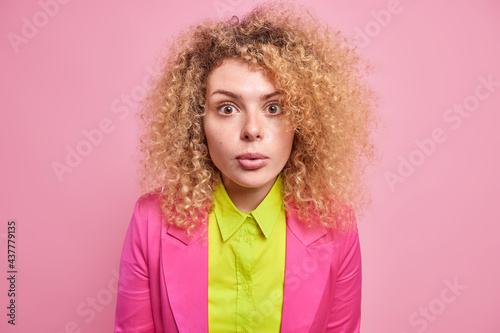 Portrait of surprised curly haired European woman stares stunned at camera hears astonishing news dressed in elegant clothes isolated over pink background cannot believe in something amazing