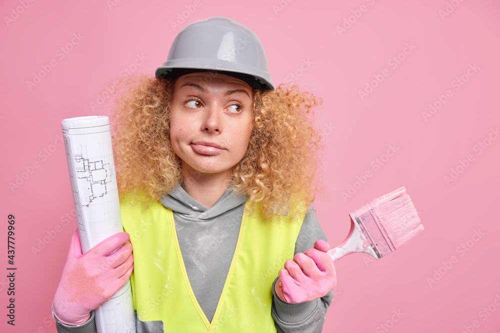 Thoughtful hesitant female builder contractor looks away with clueless expression holds painting brush and blueprint works on house remodeling has curly hair wears safety clothes hardhat gloves Thoughtful hesitant female builder contractor looks away with clueless expression holds painting brush and blueprint works on house remodeling has curly hair wears safety clothes hardhat gloves
